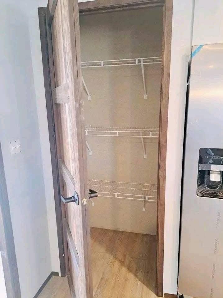 A wooden pantry door opens to reveal an empty storage space with three white wire shelves. The scene is bordered by a light wall and a stainless refrigerator.