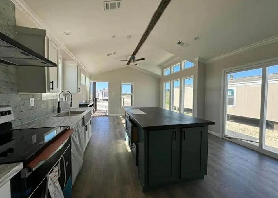 Modern kitchen interior with neutral tones. Features a central island, wood flooring, large windows with natural light, and a ceiling fan.