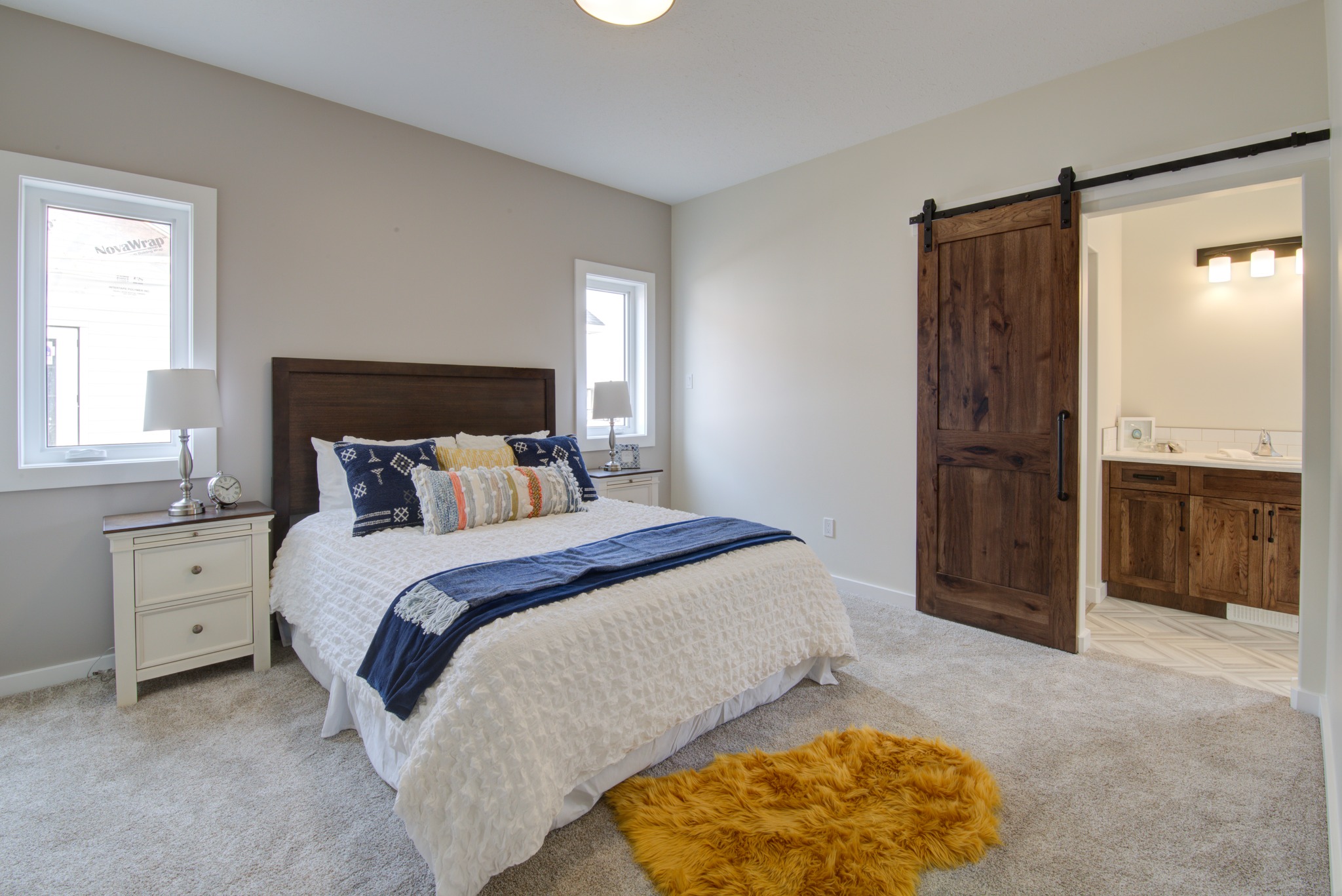 A cozy bedroom features a neatly made bed with decorative pillows, flanked by nightstands and lamps. It has a rustic sliding barn door leading to a well-lit bathroom.