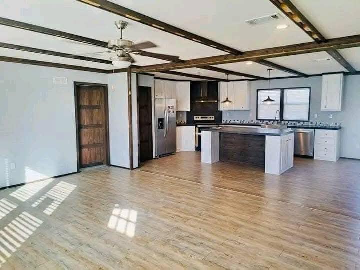 Open-concept kitchen and living area with light wood flooring and dark wood beams. Features include a ceiling fan, island, stainless steel appliances. Cozy and modern.