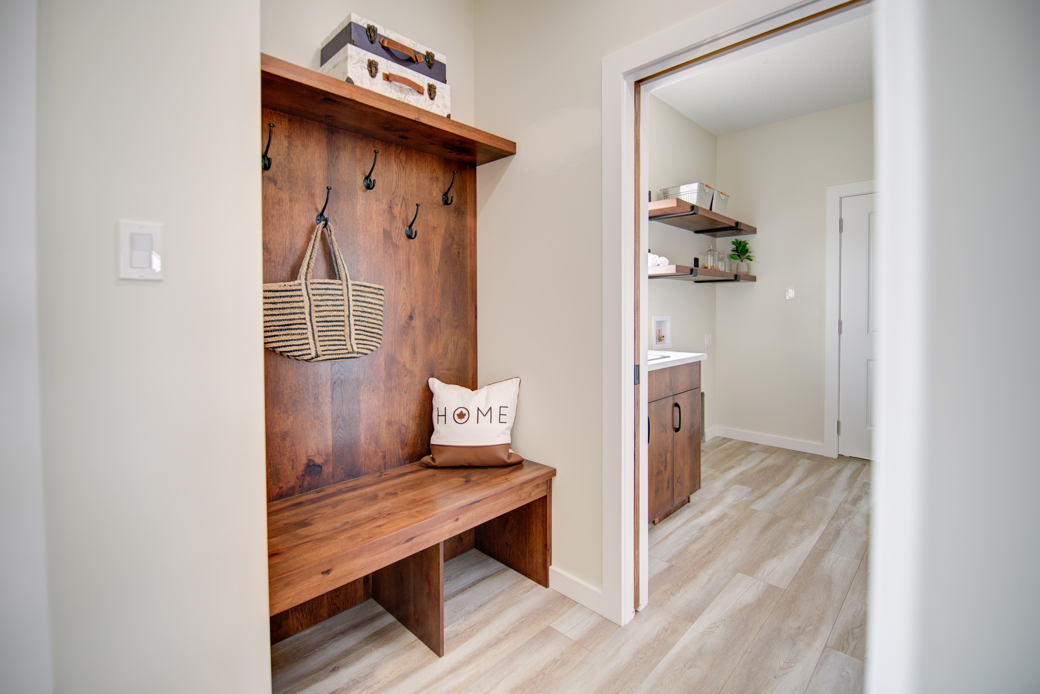 Cozy entryway with a wooden bench featuring hooks, a woven bag, and a "Home" pillow. Adjacent room has wood cabinets and light flooring.