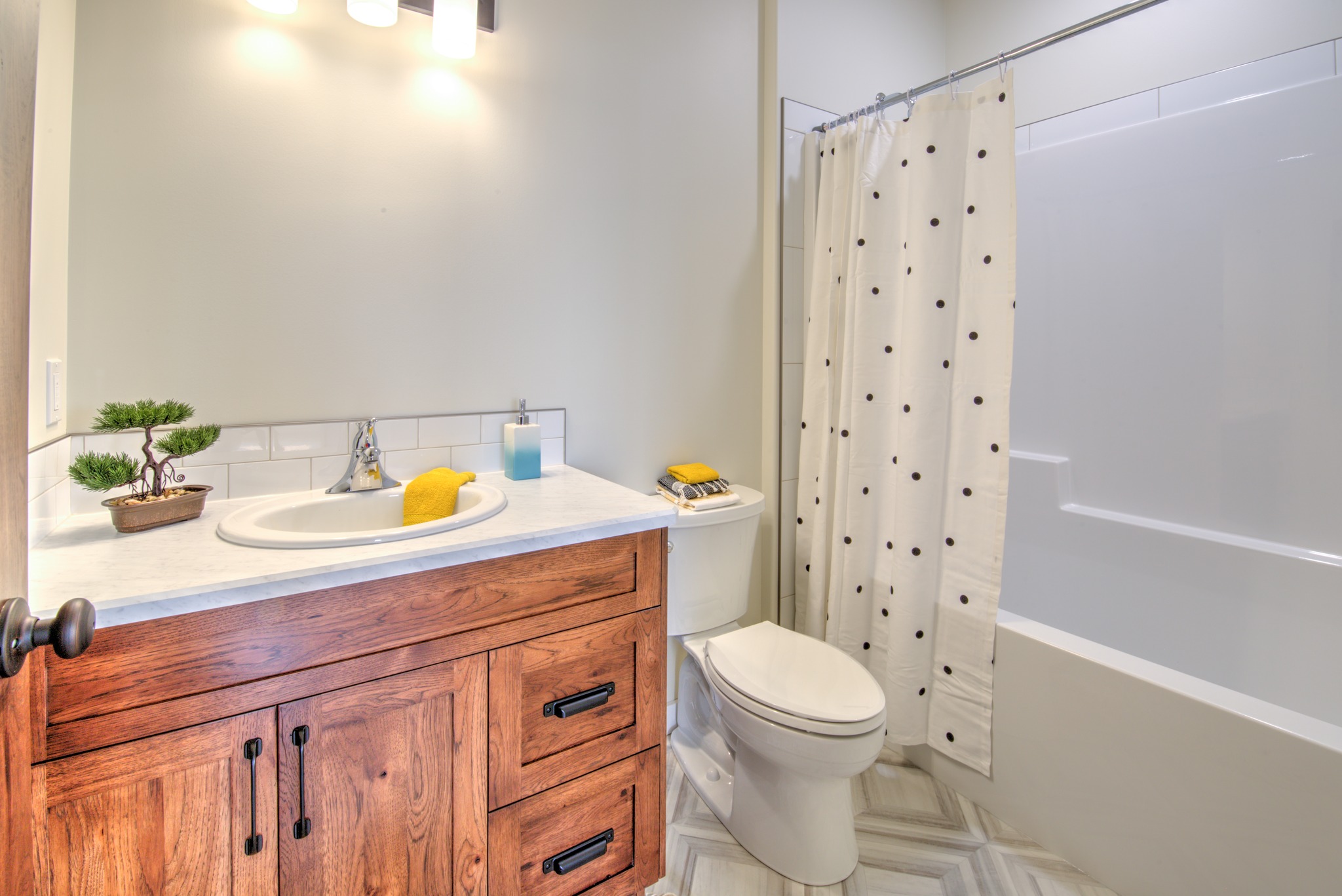 Elegant bathroom with a wooden vanity, white sink, and lit mirror. A polka dot shower curtain hangs beside a toilet in a serene, modern space.