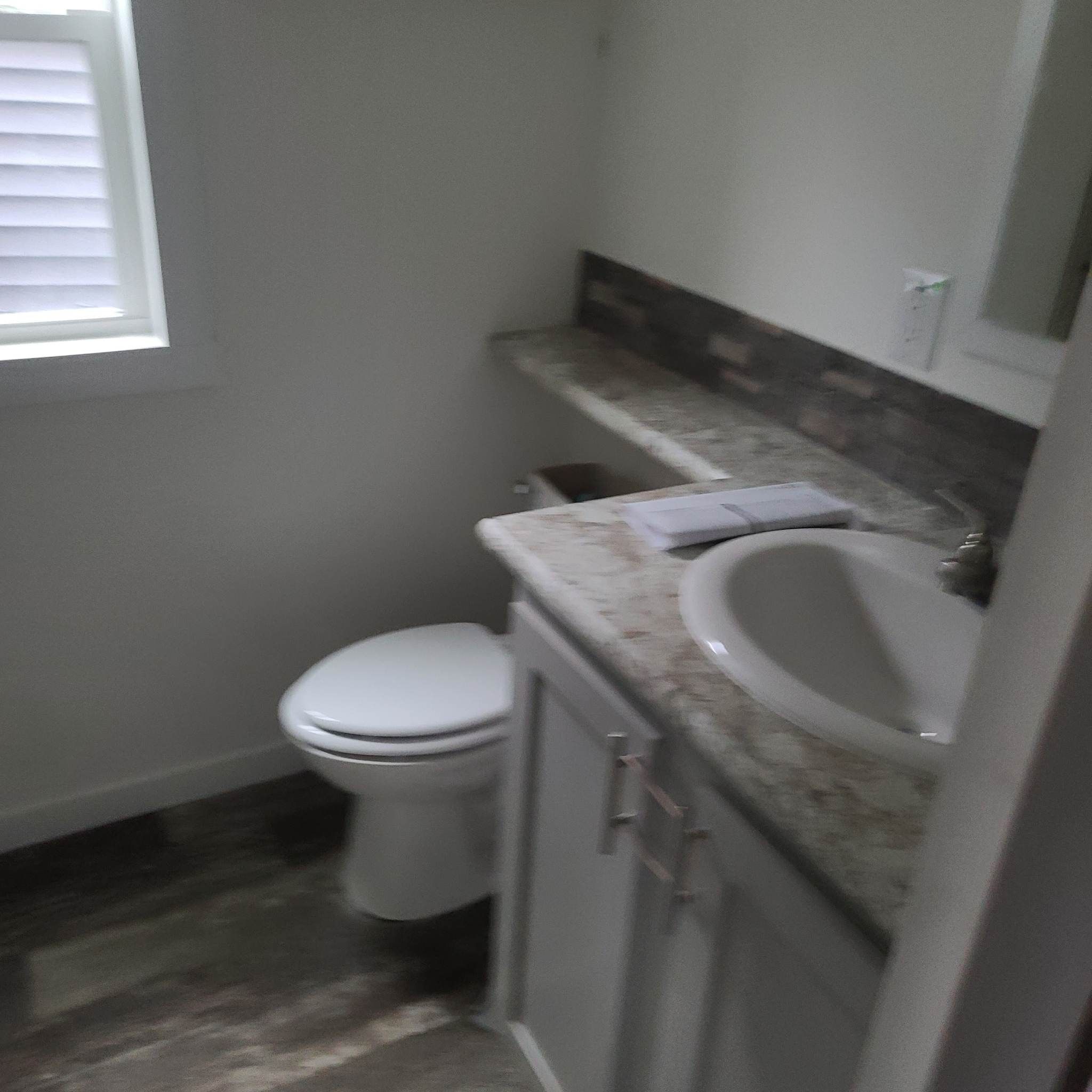 A small bathroom with a white toilet, marble sink countertop, and stone tile backsplash. On the counter is an open book. A window with blinds lets in natural light.