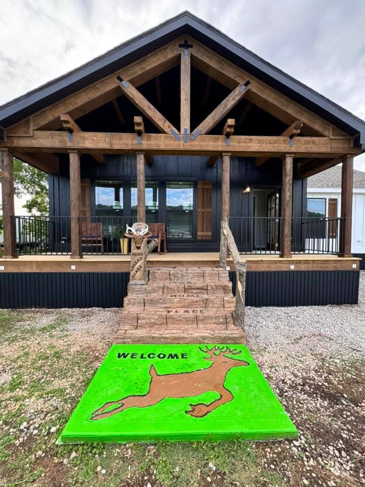 The image shows a rustic cabin with wooden beams, a welcoming front porch, and a ramp. A green mat with a deer and "WELCOME" adds charm.