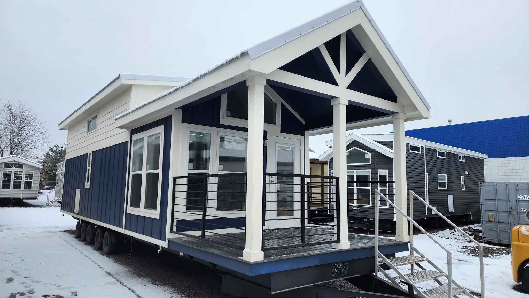 Modern tiny house with a blue and white exterior on a snowy lot. Features large windows, a small porch with black railings, and a gabled roof.