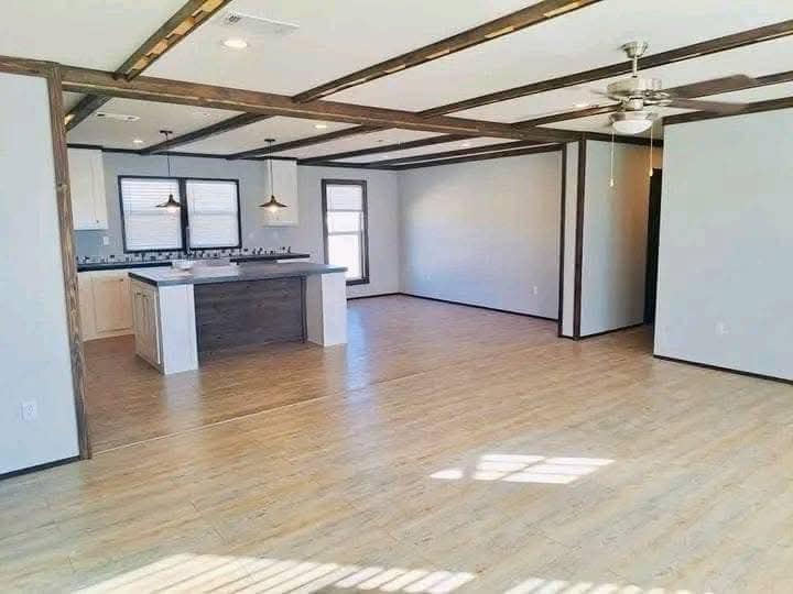 Spacious open-concept living area with light wood flooring, a modern kitchen island, pendant lights, white walls, and exposed wood ceiling beams.