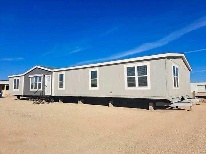 A light gray modular home on a sandy lot under a clear blue sky. The home features multiple windows and a small staircase leading to the door.