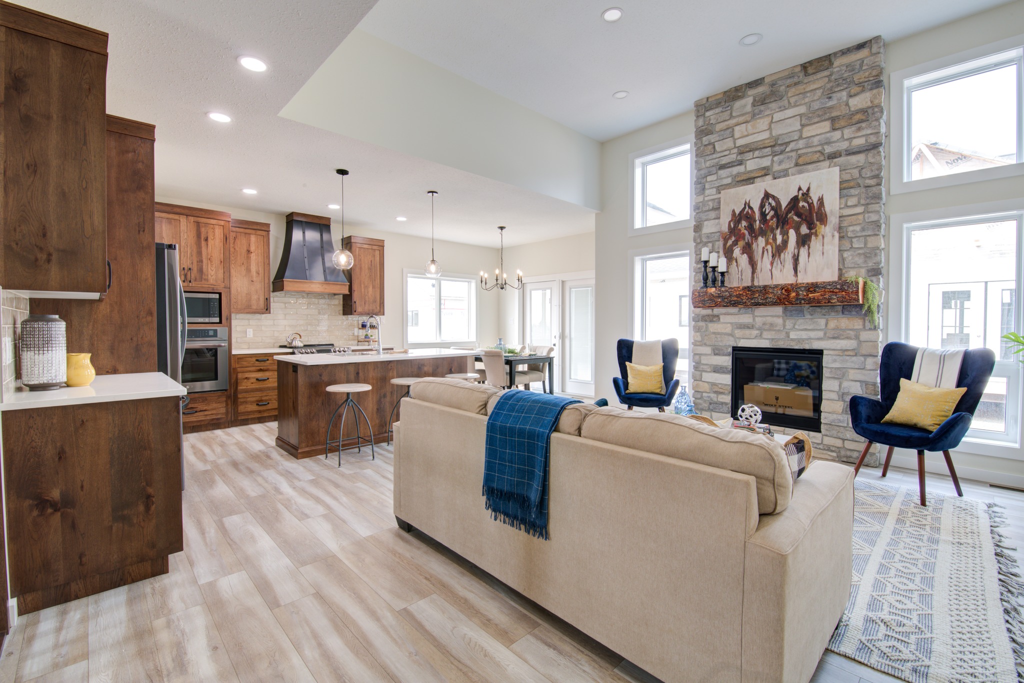 Spacious living area with a beige sofa and blue accents, adjacent to a modern kitchen. Stone fireplace, large windows, and warm wood tones create a cozy atmosphere.