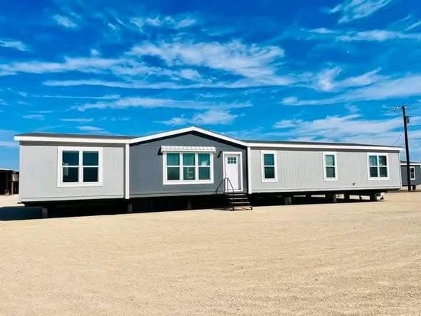 A wide, modern mobile home with white siding stands on a sandy lot under a bright blue sky with wispy clouds, conveying a sense of openness and tranquility.