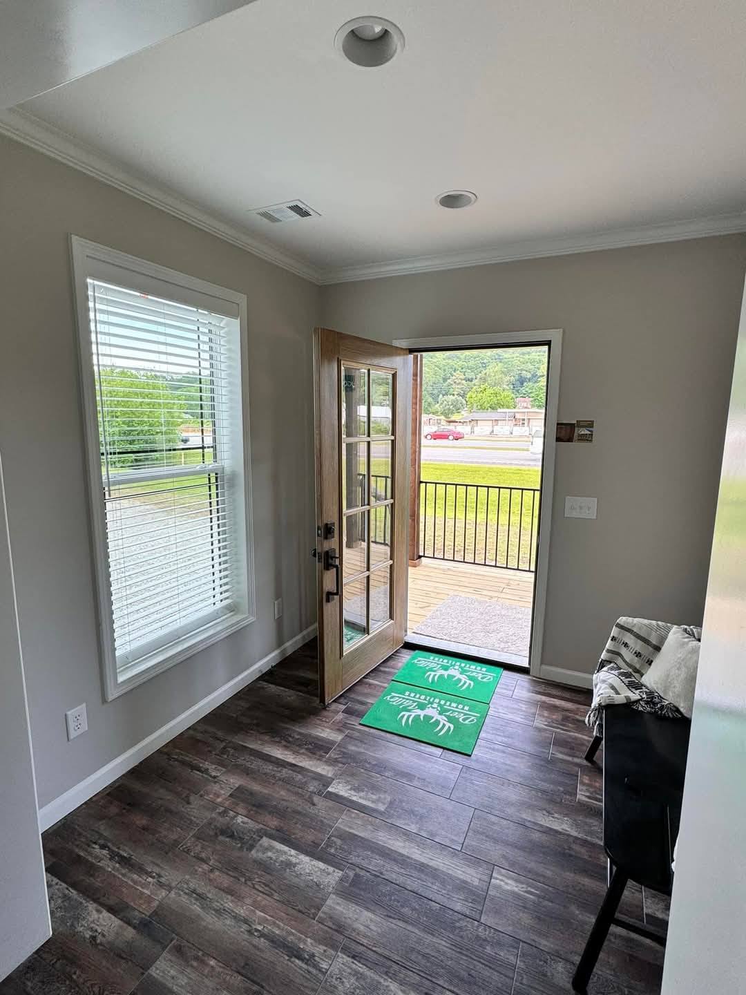 Bright entryway with wooden flooring, an open glass door leading outside. A green "Welcome" mat lies on the floor. Sunlight streams through a large window.
