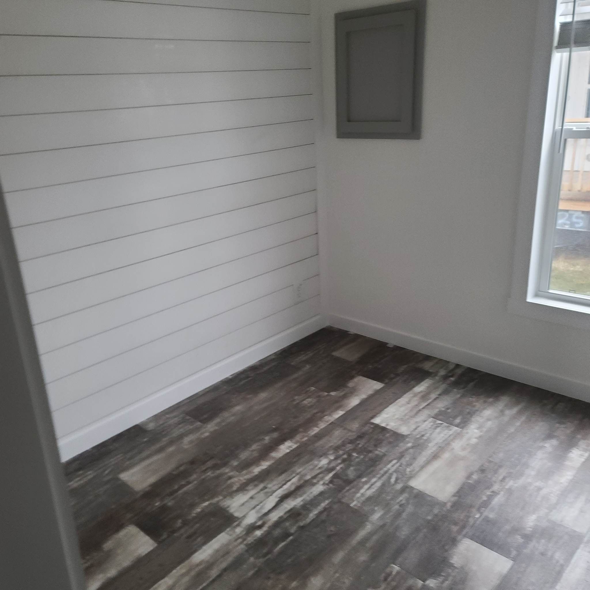 Small empty room with white shiplap walls and a single window, featuring dark wood-paneled flooring. The atmosphere is clean and minimalist.