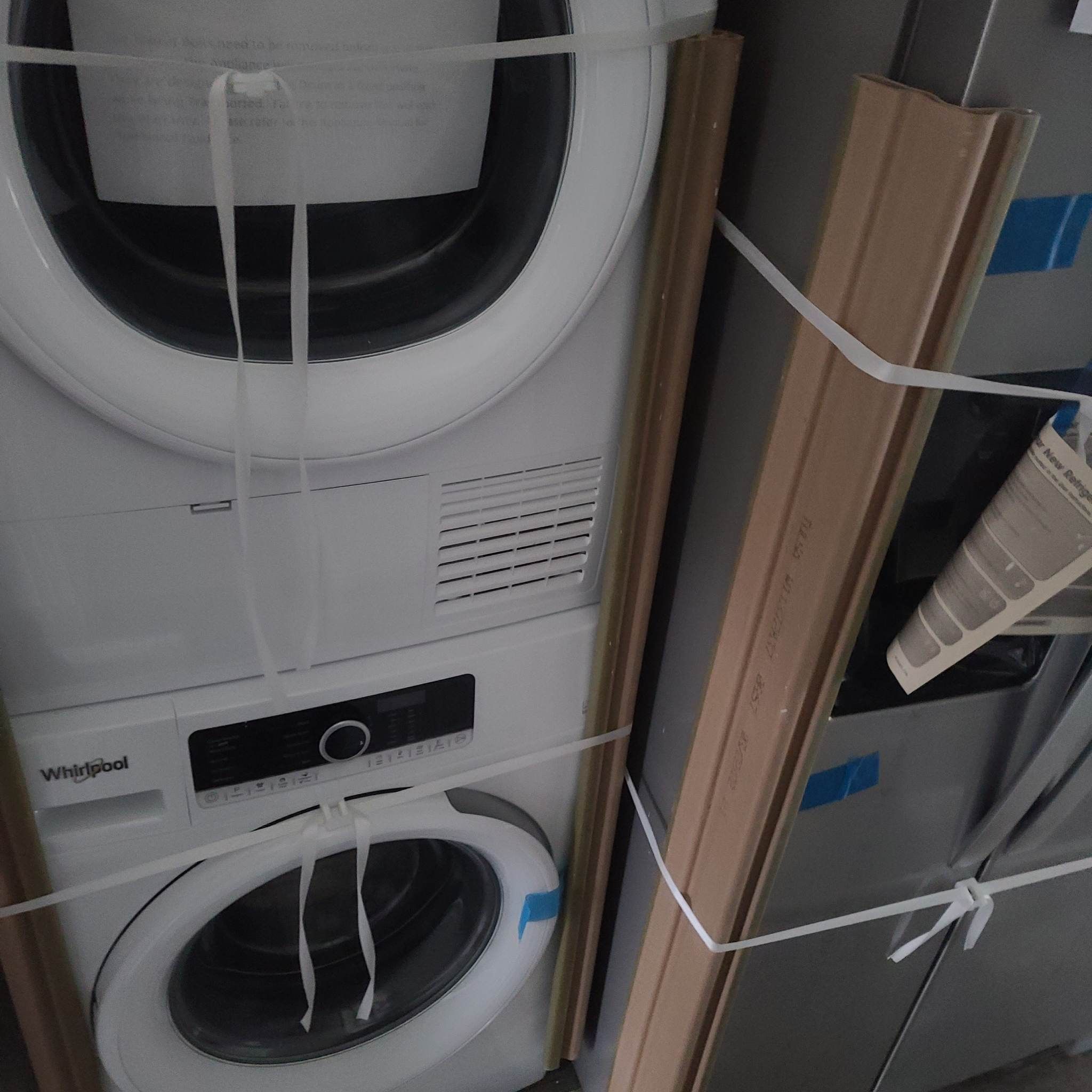 Stacked washer and dryer, secured with white straps and cardboard edges, displaying a Whirlpool logo. The setting feels utilitarian and organized.