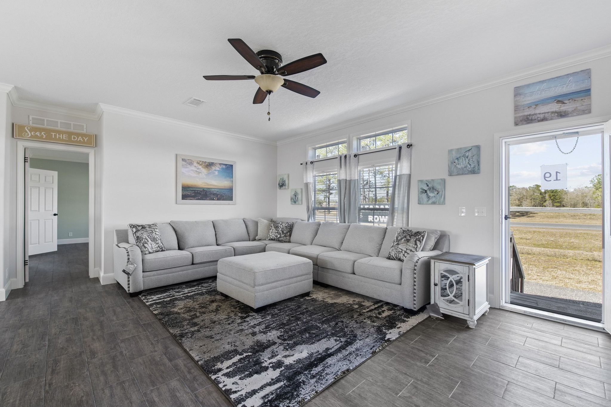 Modern living room with a large gray sectional sofa and matching ottoman on a textured rug. White walls with beach-themed artwork, wood floor, and windows bring in natural light. A ceiling fan and a sign reading "Seas the Day" add charm.