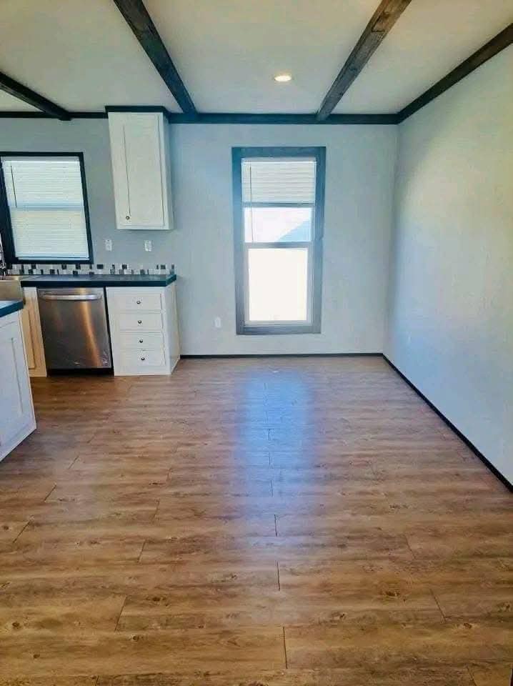A bright room with wood flooring, white walls, and ceiling beams. On the left, a kitchen area features white cabinets and a stainless steel dishwasher.