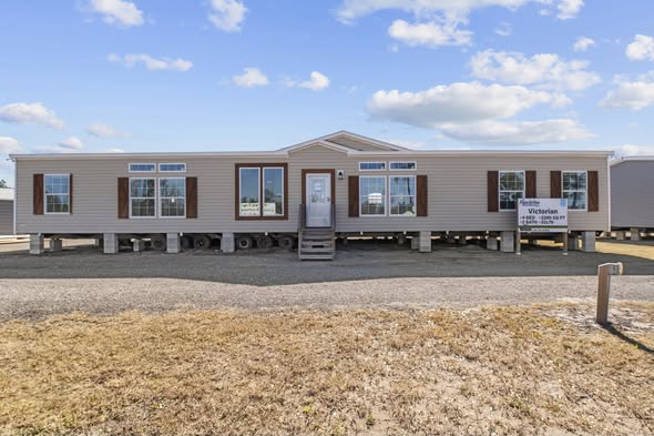 A beige modular home elevated on stilts with brown shutters against a clear blue sky. A small sign reads "Victorian" near the entrance, evoking a sense of new beginnings and stability.