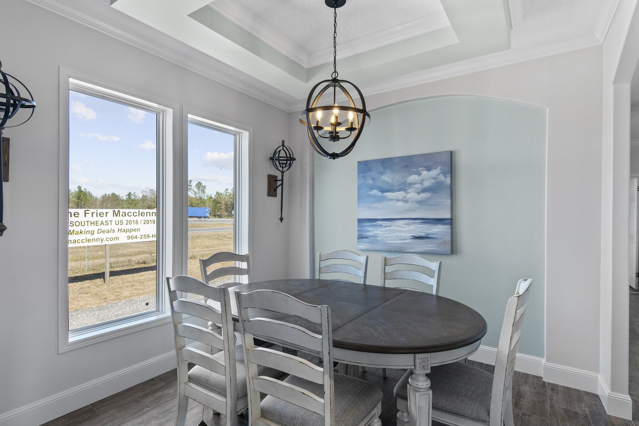 Dining room with elegant chandelier, wooden table, and six chairs. Large windows offer natural light. Ocean painting on a blue accent wall. Airy and serene.