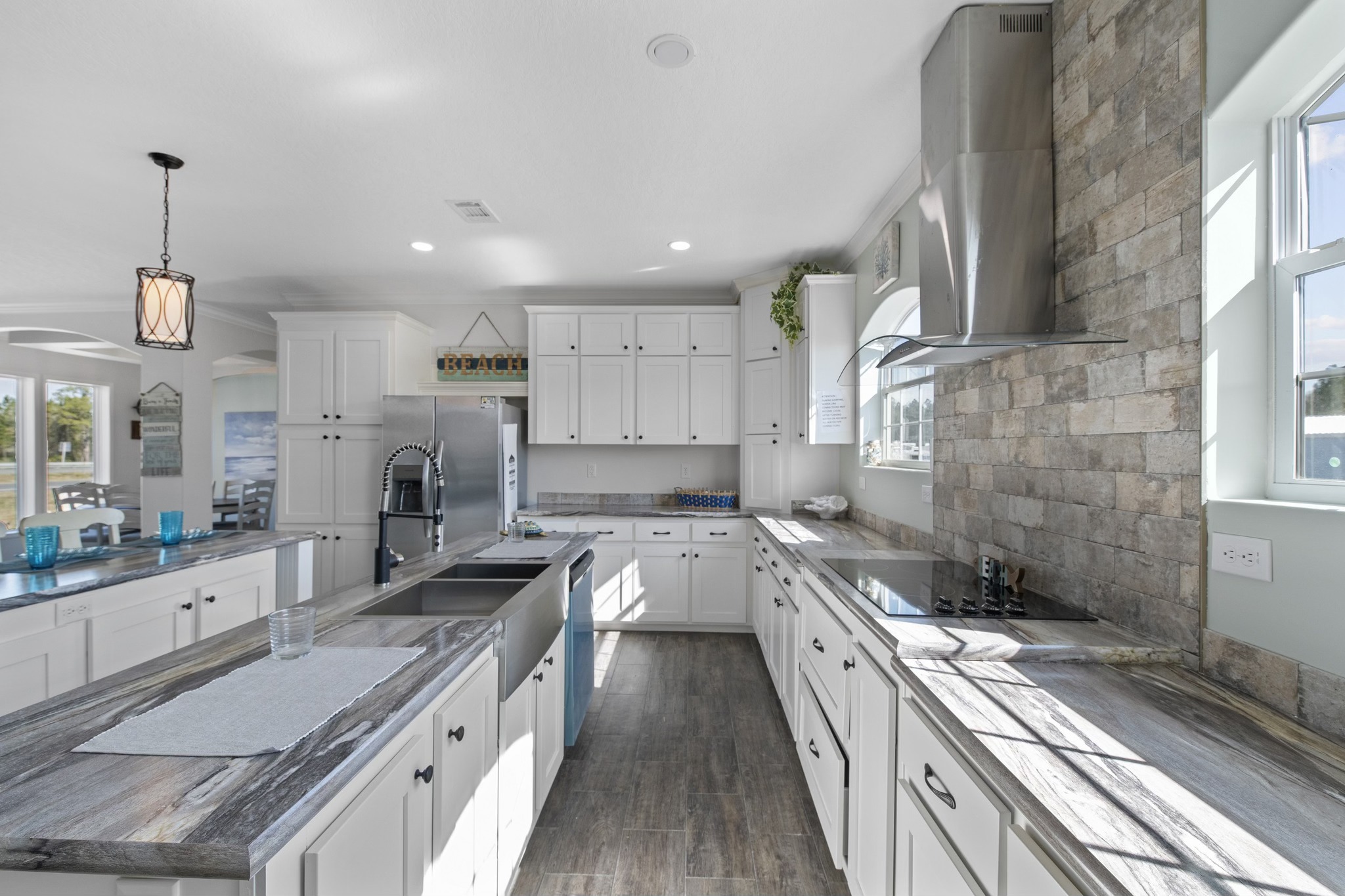 Bright, modern kitchen with white cabinets, stainless steel appliances, stone backsplash, and large windows. Sunlight casts soft shadows on the countertops.