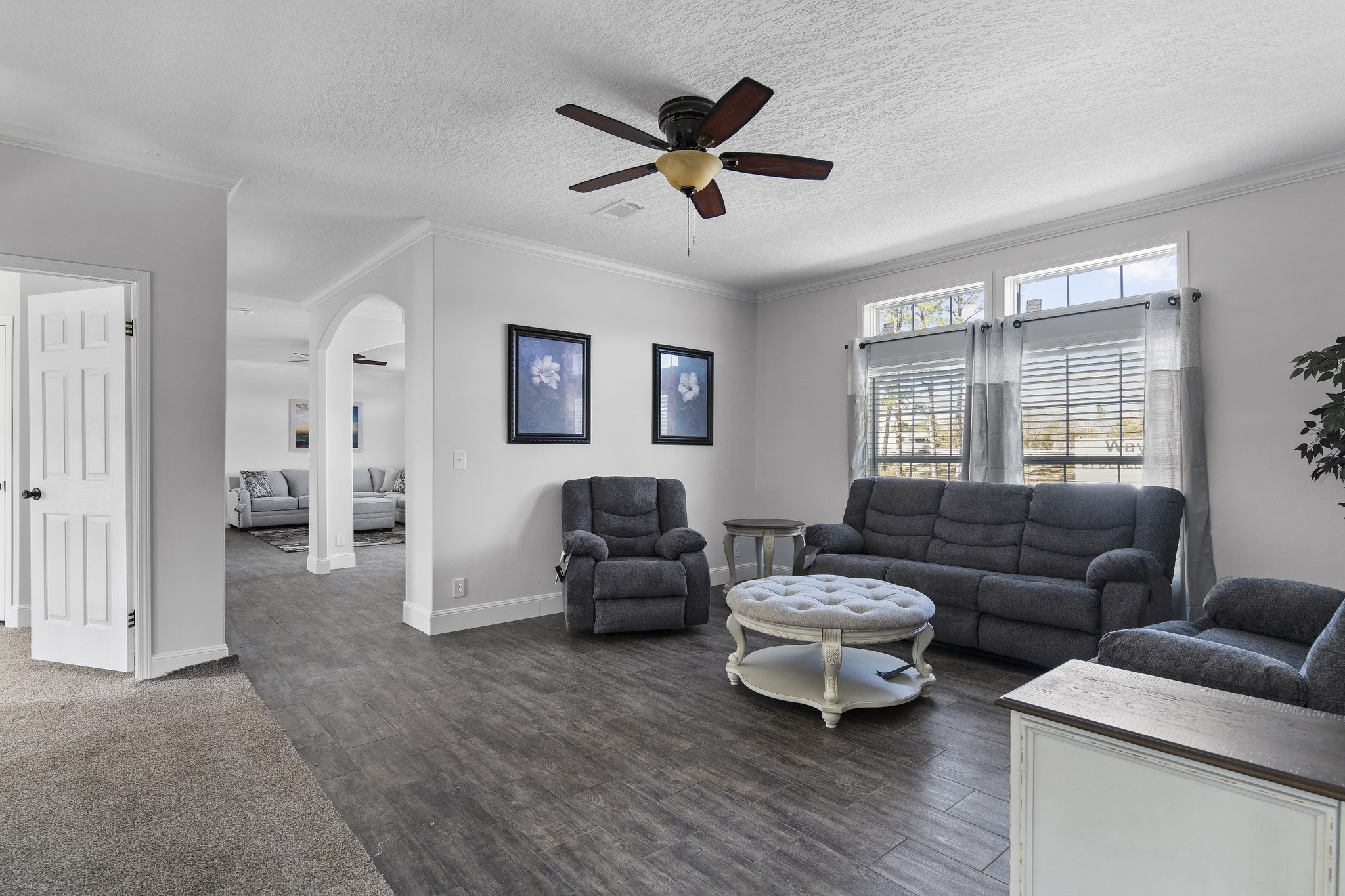 A cozy living room with gray sofas and armchairs, a white round coffee table, and a ceiling fan. Sunlight streams through curtained windows, creating a serene ambiance.