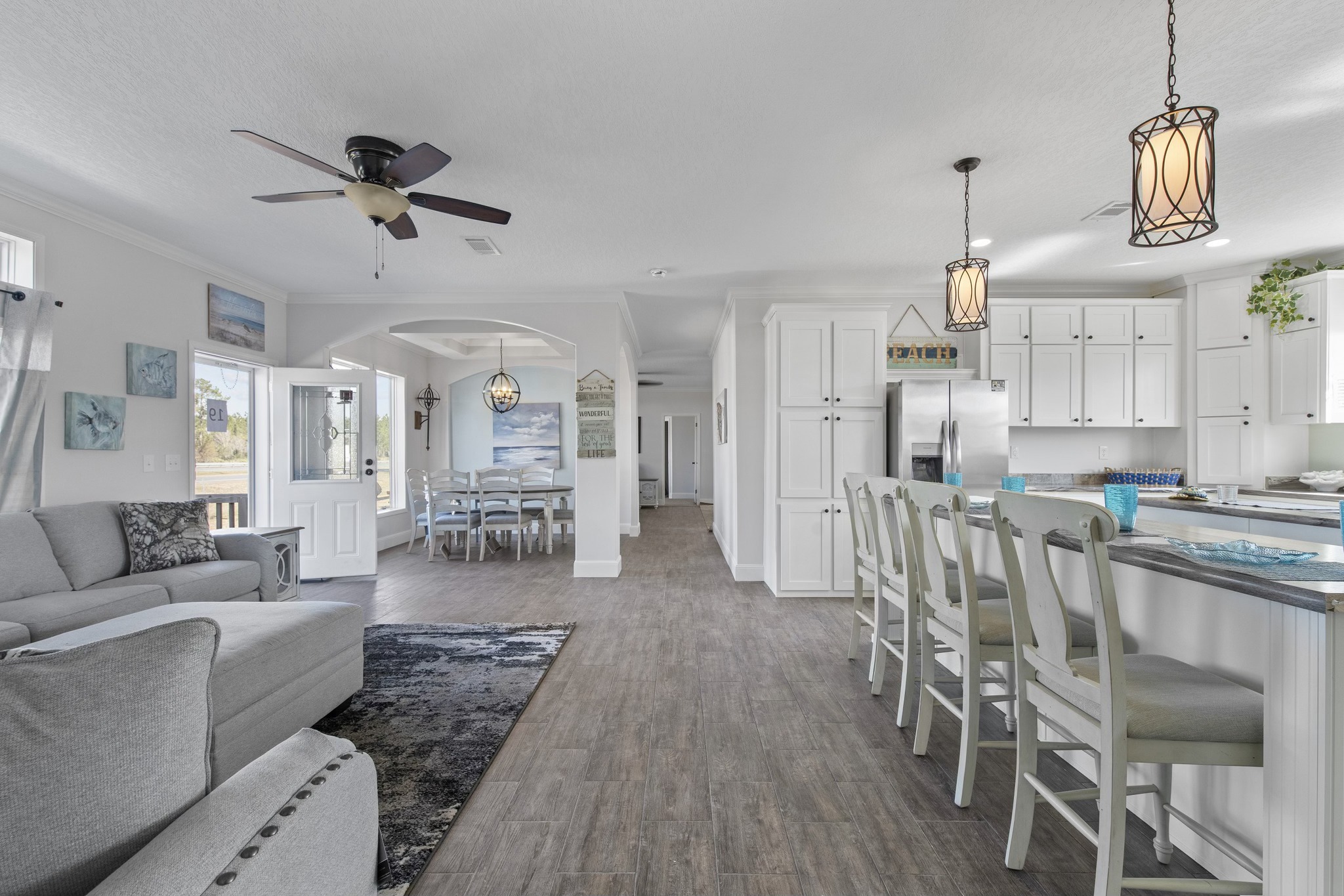 Open-concept living room and kitchen with modern coastal decor. Features gray sofa, dining area, white cabinetry, bar stools, and pendant lights.