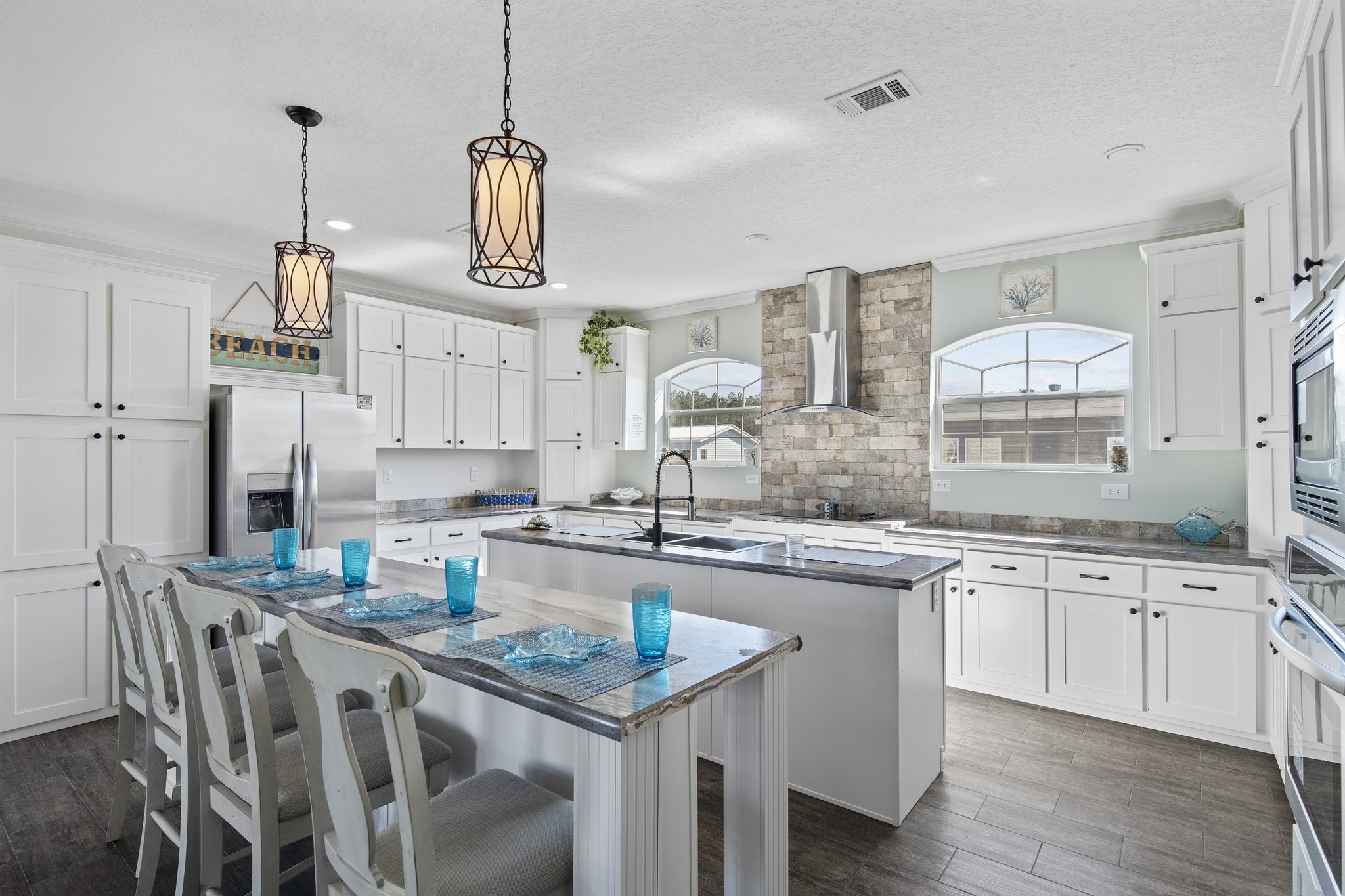 Spacious, modern kitchen with white cabinets, island countertop, pendant lights, and bar stools. Bright atmosphere with large windows and a stainless-steel fridge.
