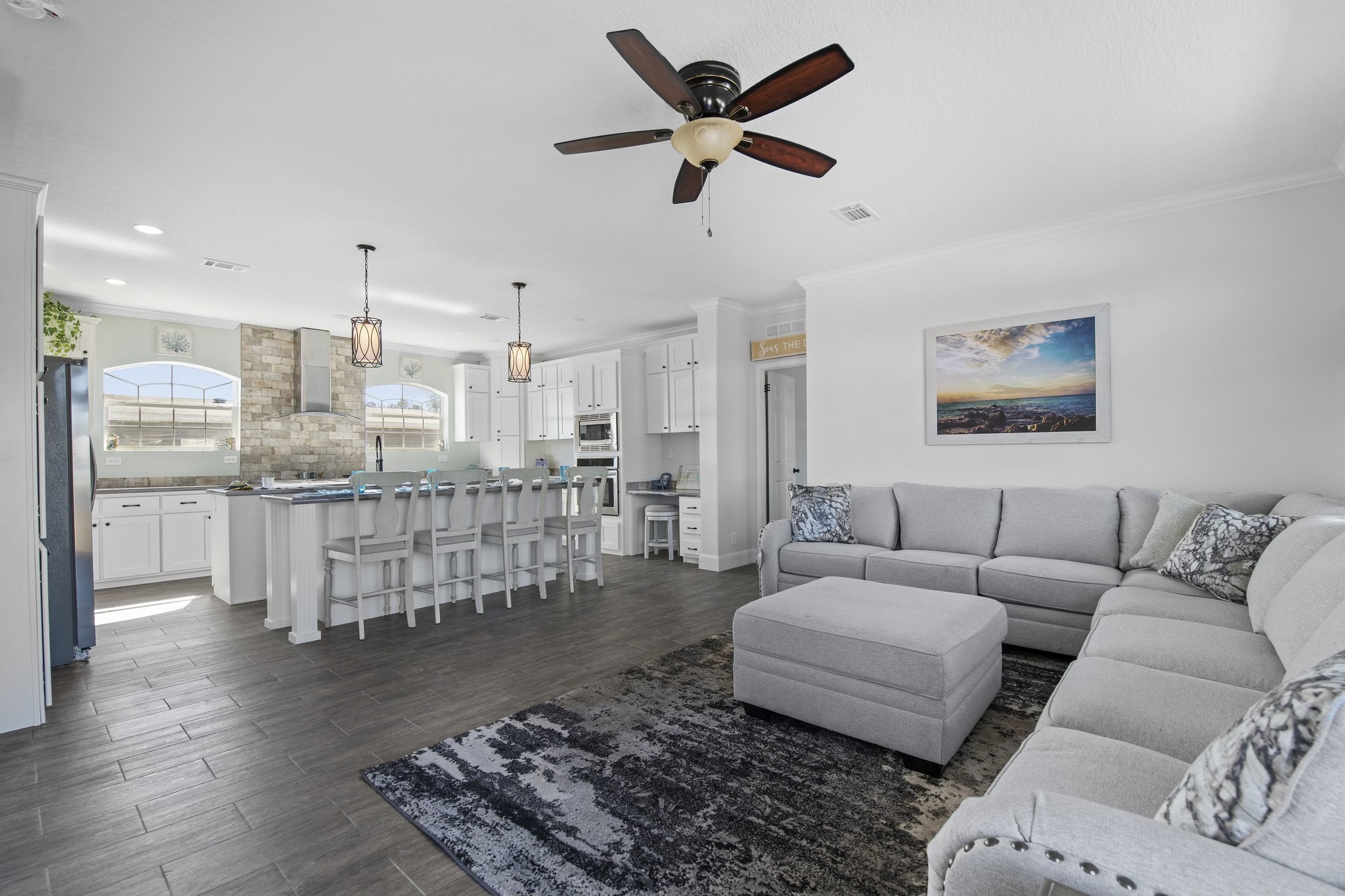 Spacious open-concept living room and kitchen, featuring a gray sectional sofa, dark wood floor, ceiling fan, and kitchen island with white barstools.