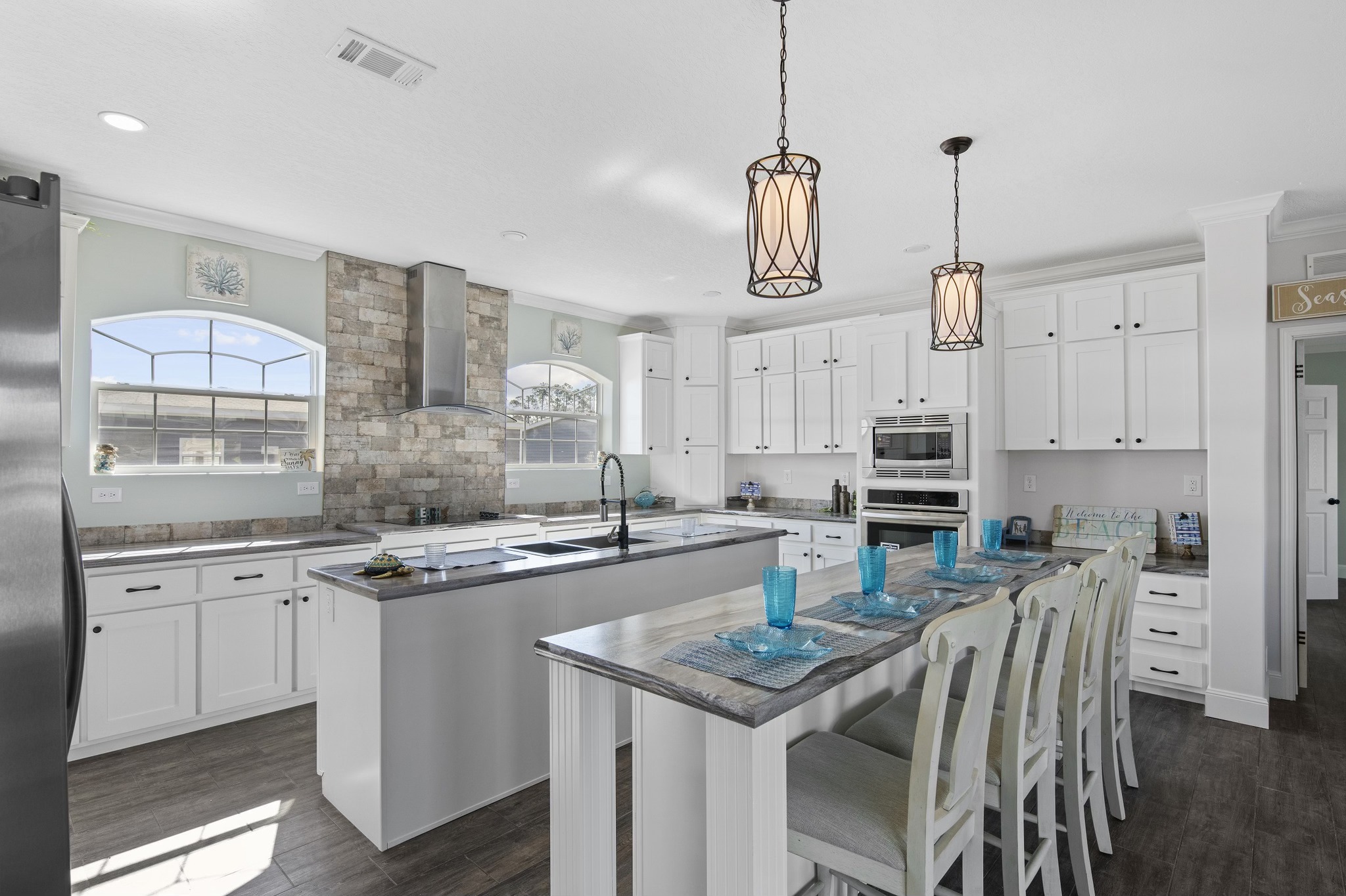 A bright, modern kitchen with white cabinetry, a stone backsplash, large island, and four high chairs. Blue glassware adds color. Warm lighting from pendant lamps.