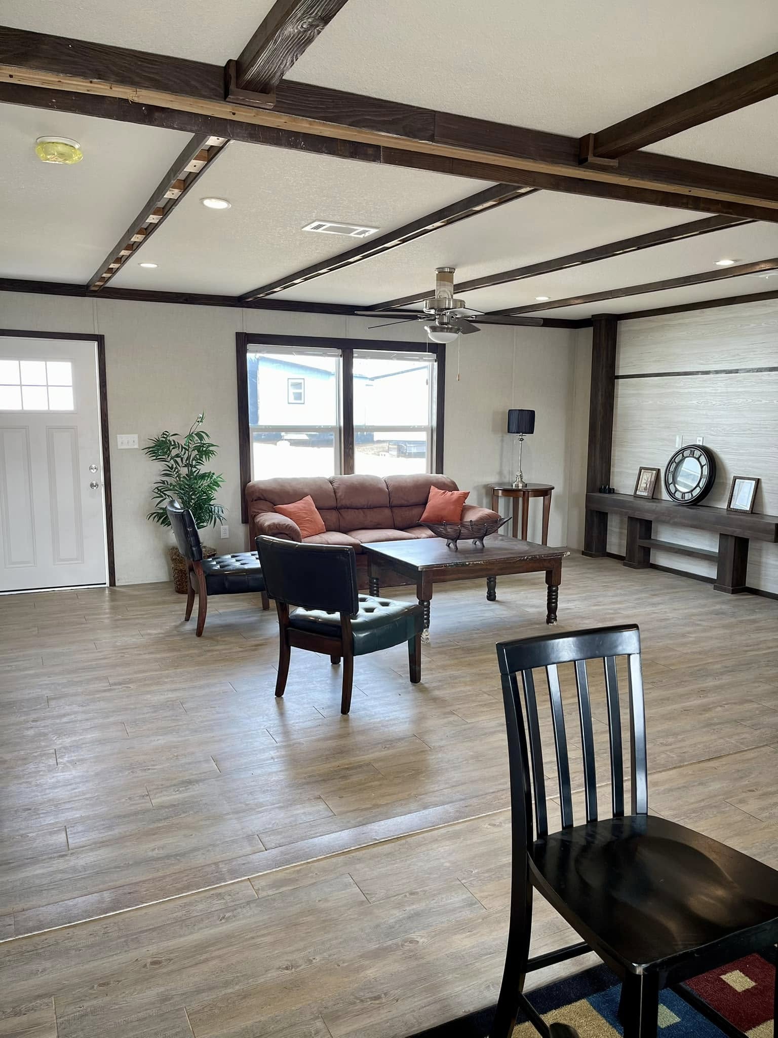 Spacious living room with a brown leather sofa, wooden coffee table, and elegant chairs. Warm tones, large window, and ceiling beams create a cozy feel.
