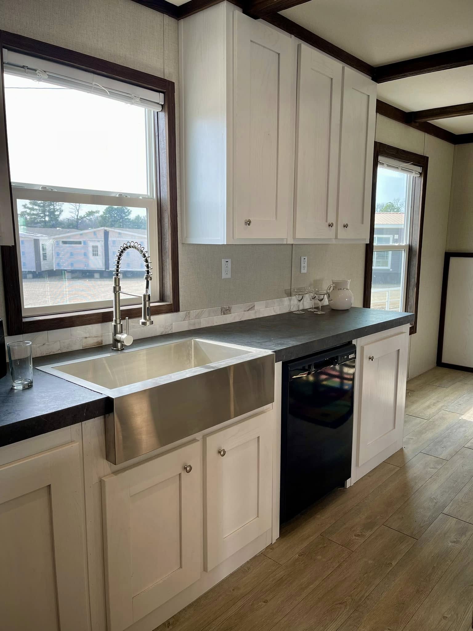 Modern kitchen with a large stainless steel farmhouse sink, black countertop, and white cabinets. Sunlight streams through two large windows.