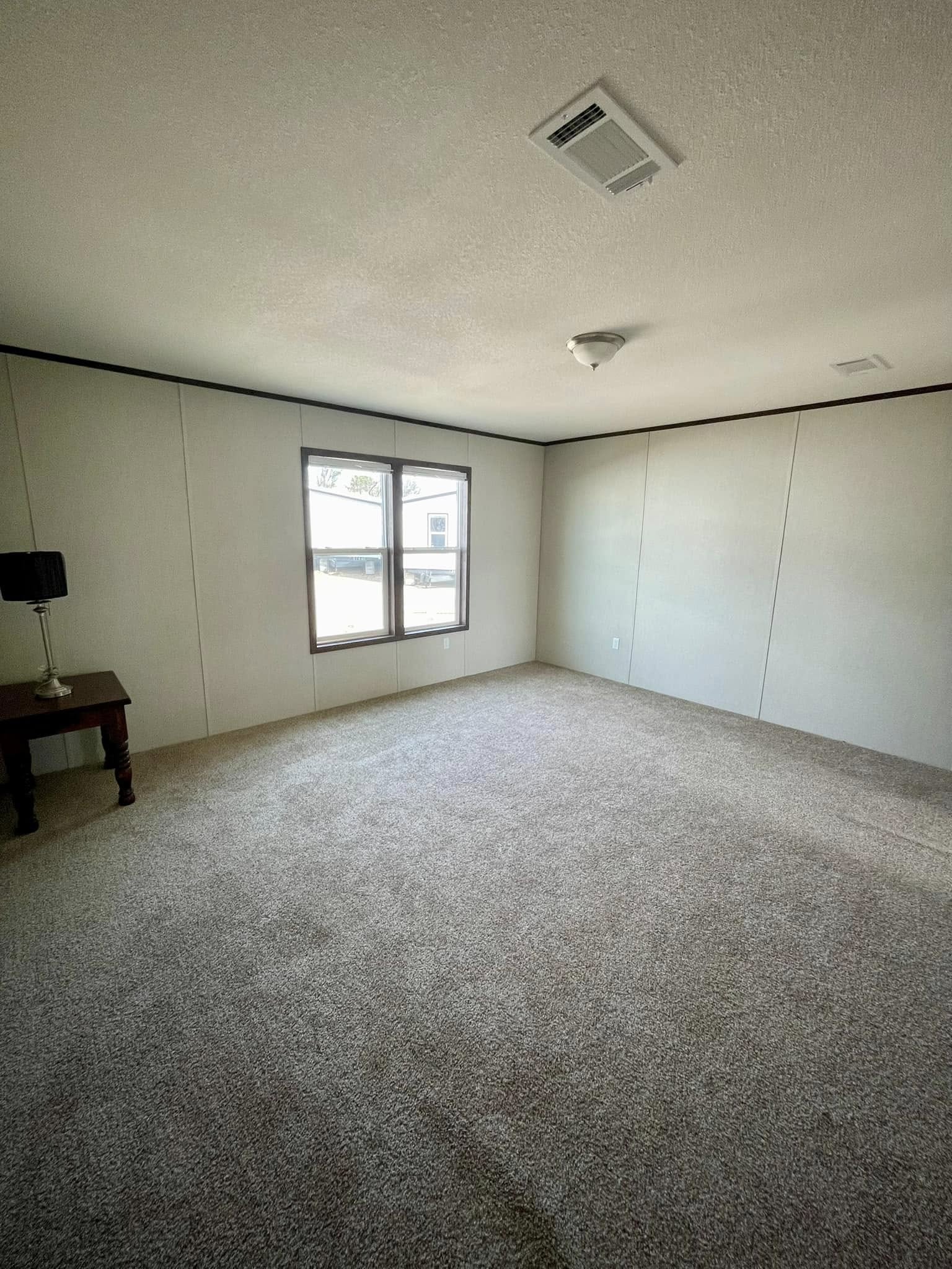 Empty room with beige carpet, white walls, and a central ceiling light. A single window allows natural light. There's a small table with a lamp.