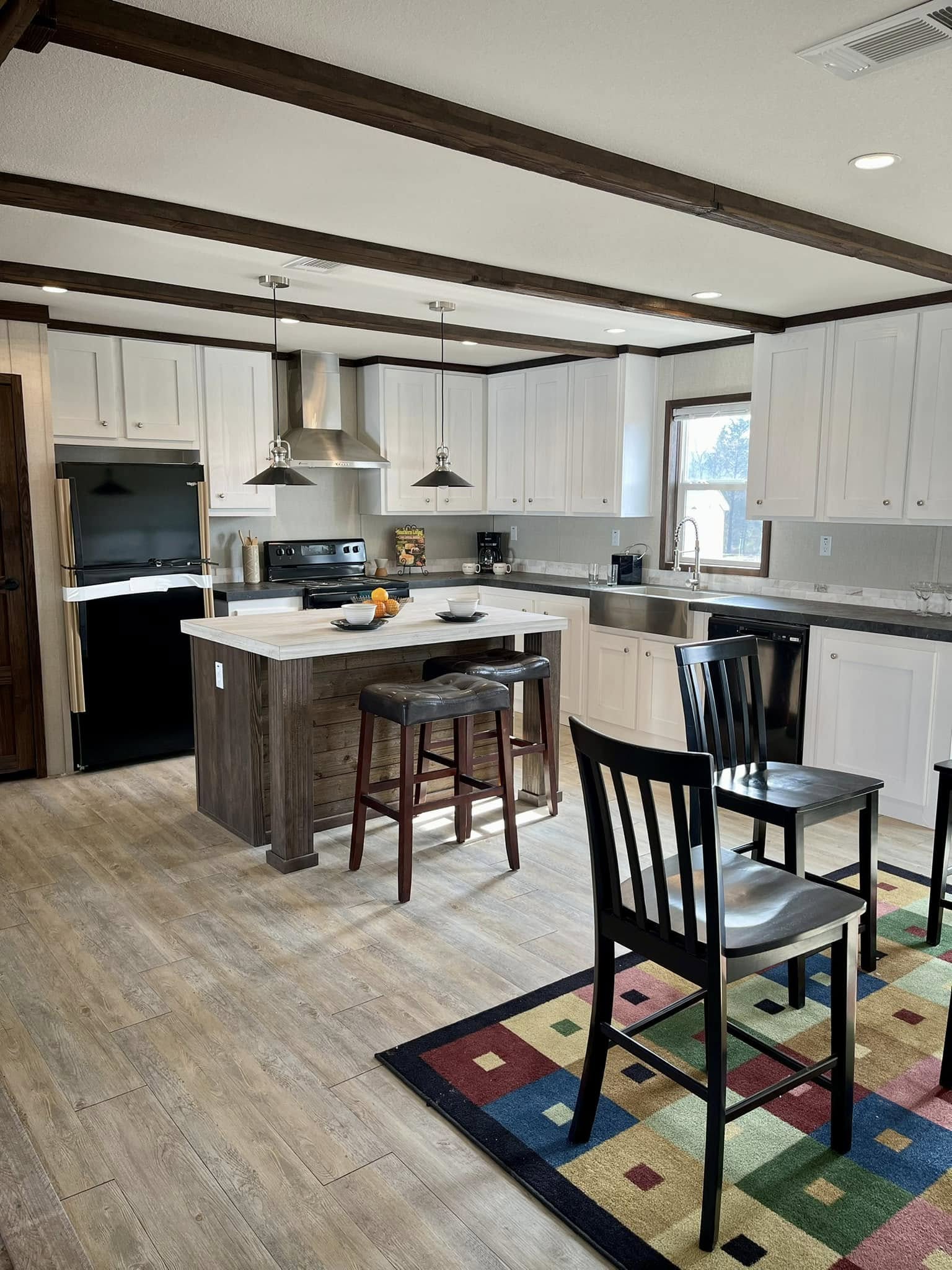Modern kitchen with white cabinetry and dark wood accents. Central island with bar stools, pendant lights, and a colorful patterned rug on wood floors. Bright, inviting ambiance with natural light from a window.