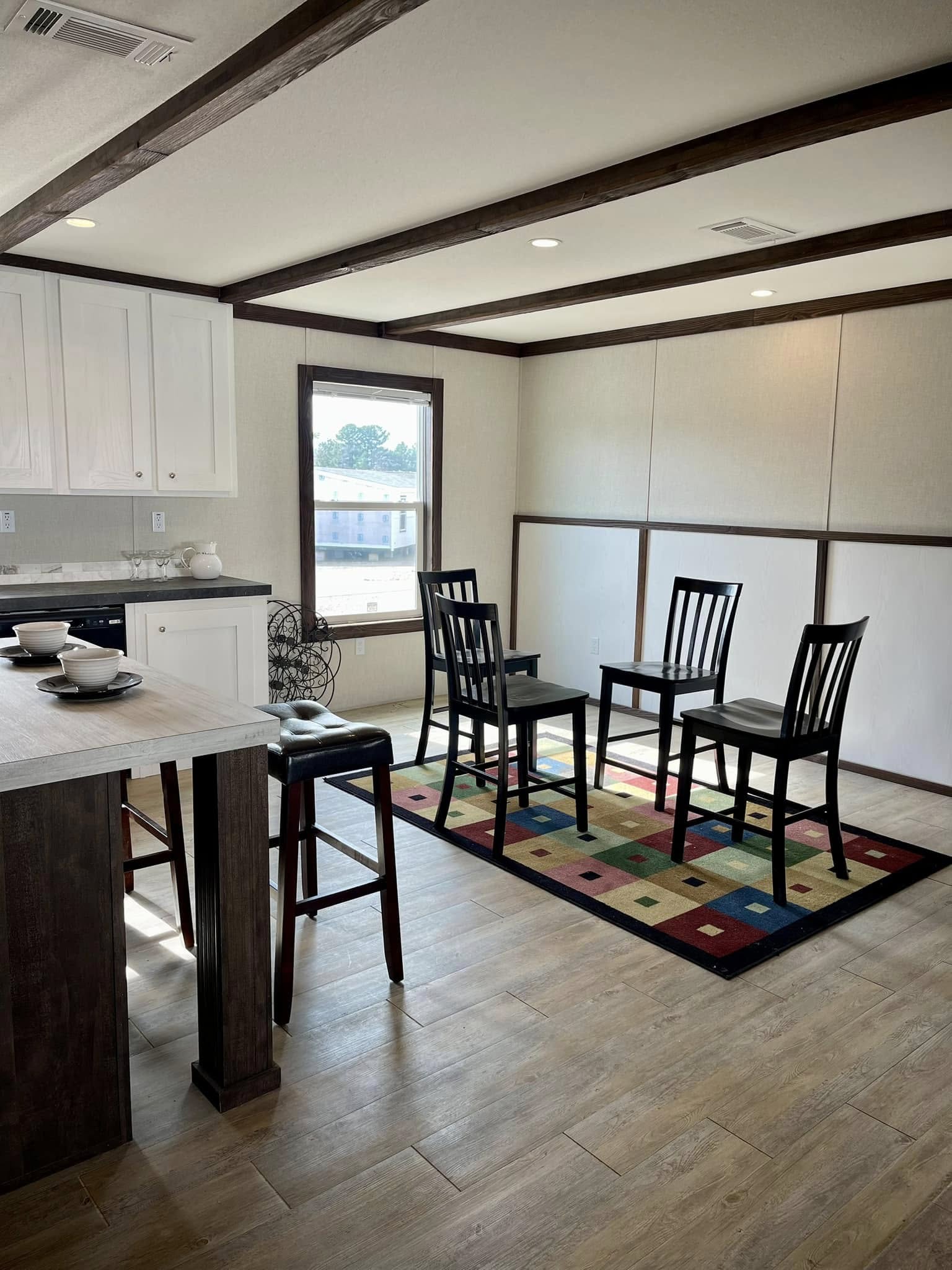 Modern kitchen with wooden floor and beams, featuring four black chairs on a colorful checkered rug. White cabinets and a breakfast bar add a cozy feel.