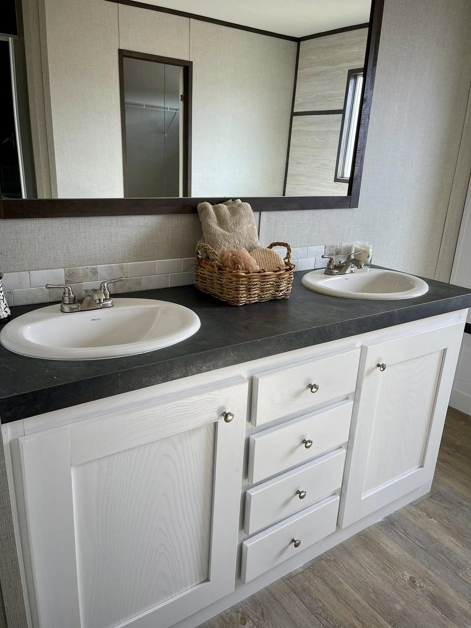 A bathroom features a double sink vanity with dark countertop and white cabinets. A wicker basket with towels and a sponge sits between the sinks.