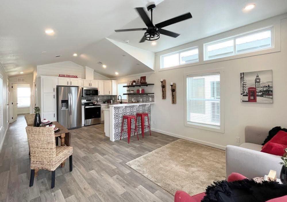 Modern living room and kitchen with light wood flooring. Features a ceiling fan, white cabinetry, stainless steel appliances, and a patterned bar with red stools. Cozy seating area with a gray sofa, wall art, and bright windows.