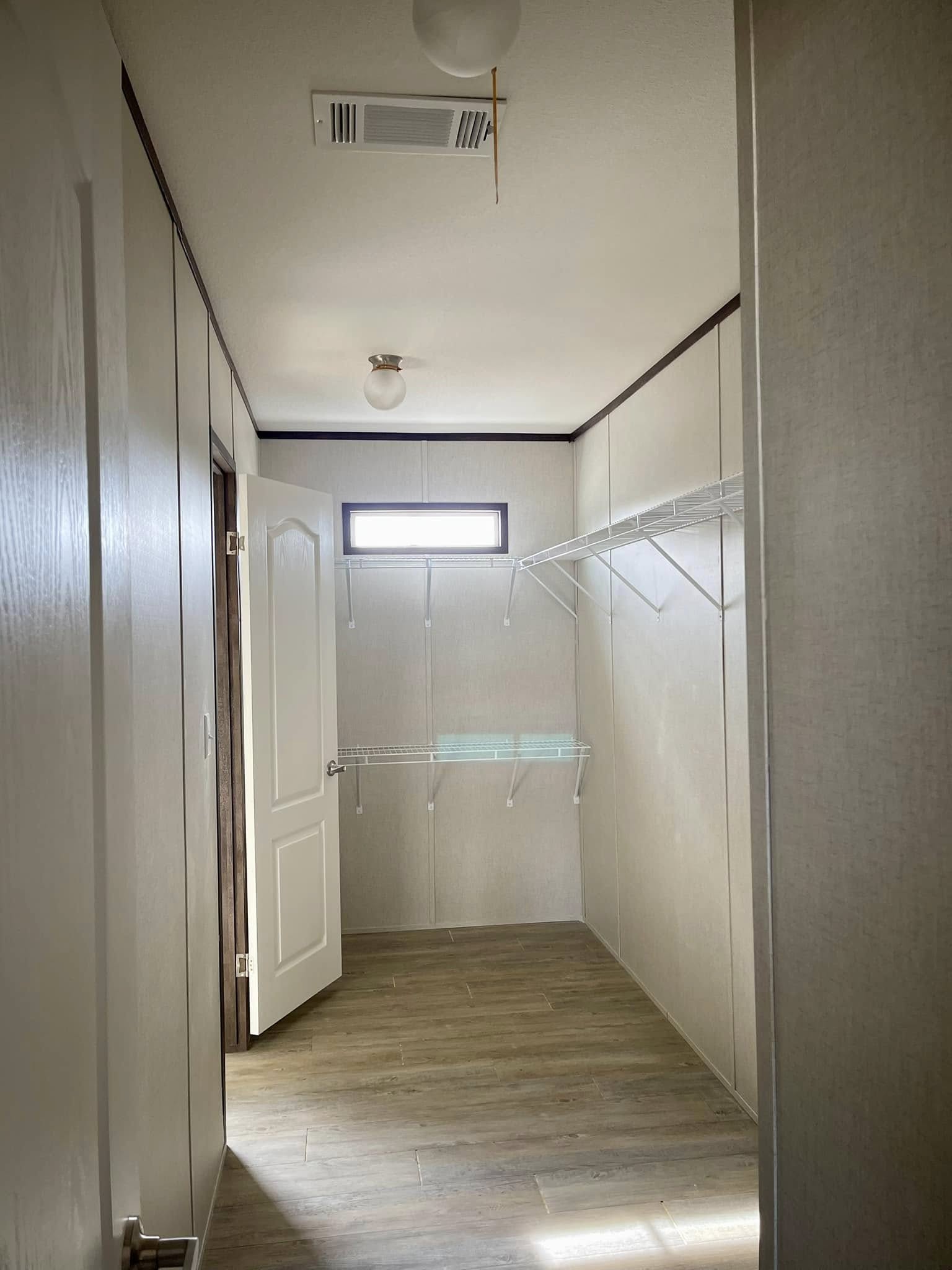 Narrow walk-in closet with light wood flooring, open white door, high window, and metal wire shelves on beige walls, creating a minimalist feel.