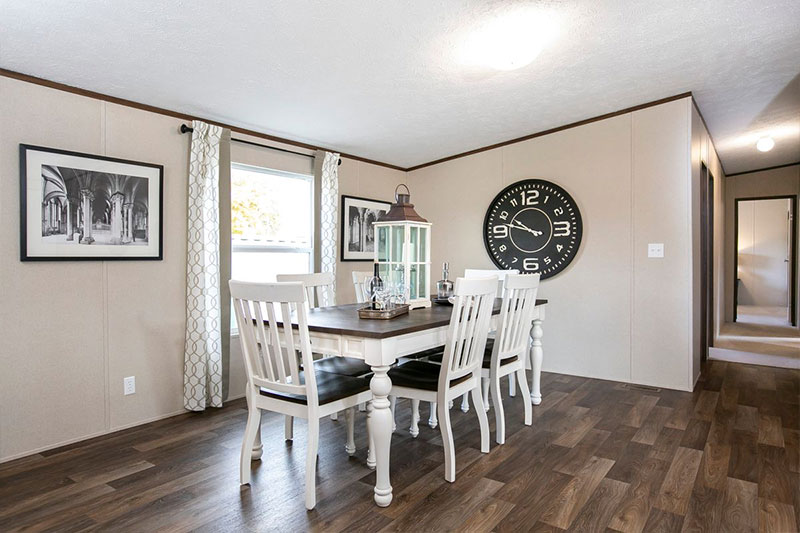 A cozy dining room with a wooden table and six white chairs. A large black clock and framed photos adorn beige walls. Warm lighting creates an inviting atmosphere.