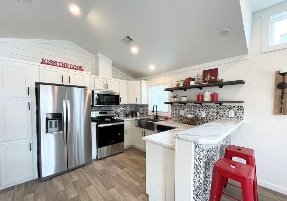 A modern kitchen with white cabinets, stainless steel appliances, and decorative black and white tile backsplash. Red accents, shelves with decor, and two red stools add a vibrant touch. The phrase "Kiss the Cook" is displayed above the cabinets.