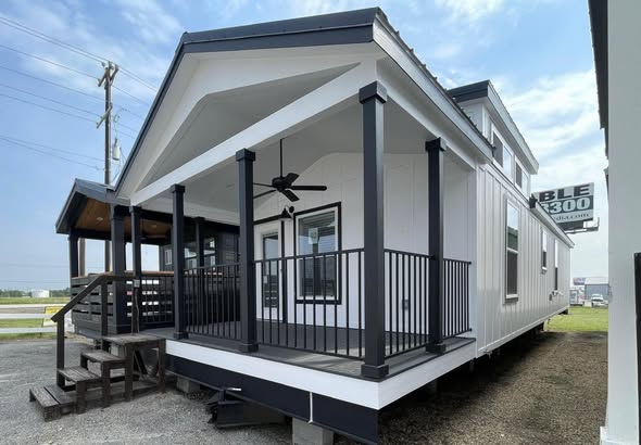 Modern tiny house with a black and white exterior, featuring a covered porch, railings, and ceiling fan. Bright sky, open ambiance.