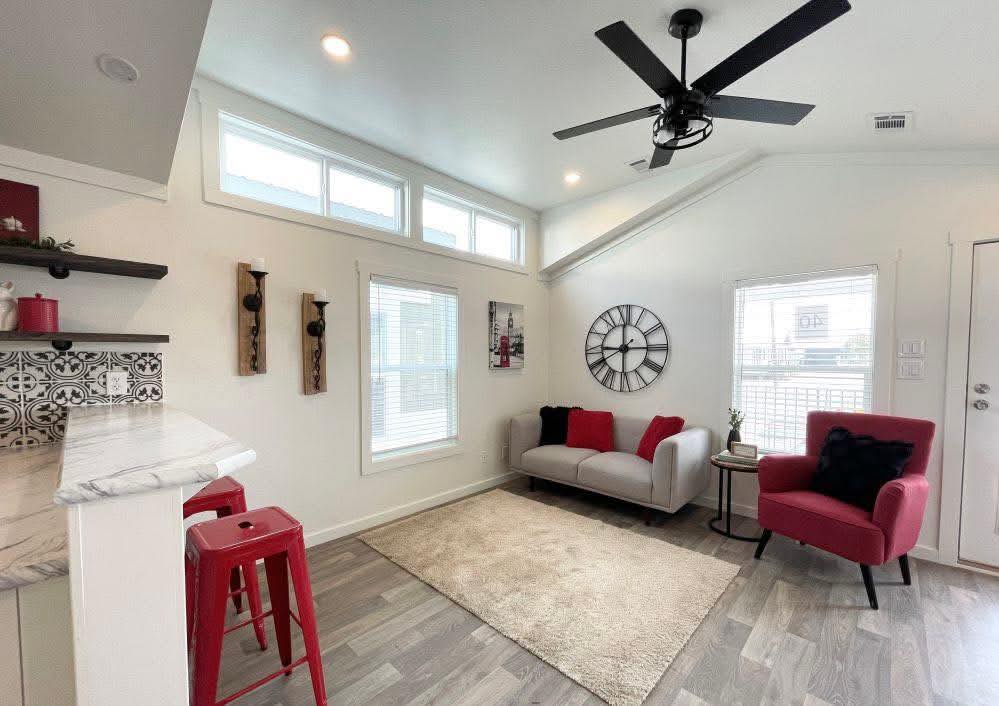 A modern living room with white walls, a light gray sofa, red chair, and two red stools. A geometric rug, large wall clock, and ceiling fan add style.