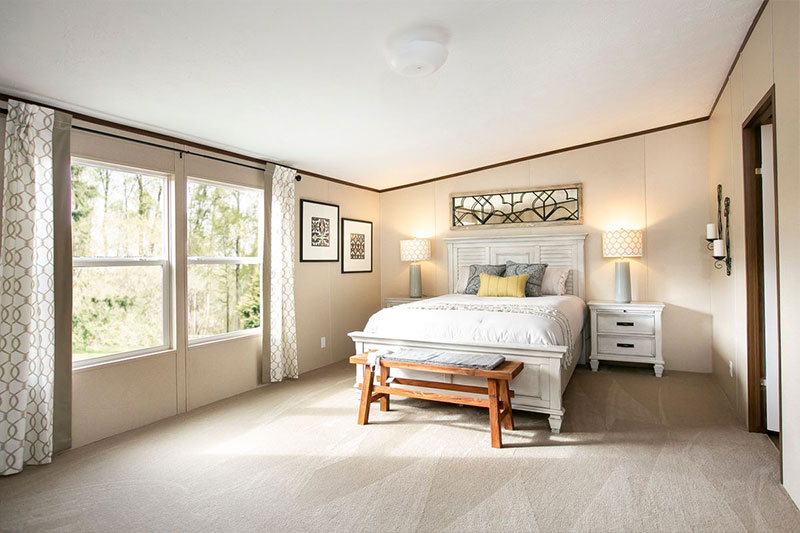 Bright bedroom with a large bed, white bedding, and a wooden bench at the foot. Two windows with patterned curtains, lamps, and light wall decor create a cozy atmosphere.