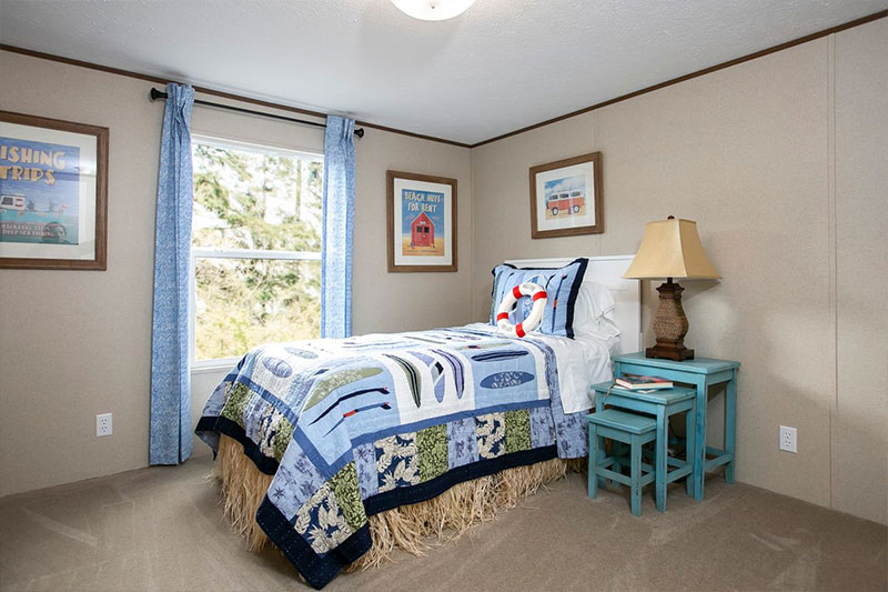 Cozy bedroom with a nautical theme, featuring a twin bed with a blue patchwork quilt. Light blue curtains frame the window, and beach-themed art adorns the beige walls. A small lamp sits on turquoise side tables, creating a serene, coastal vibe.