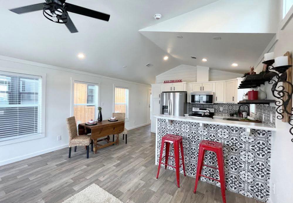Bright open-plan kitchen and dining area with white walls, patterned tile, red stools, wicker chairs at a wooden table, and large windows. Modern and inviting.