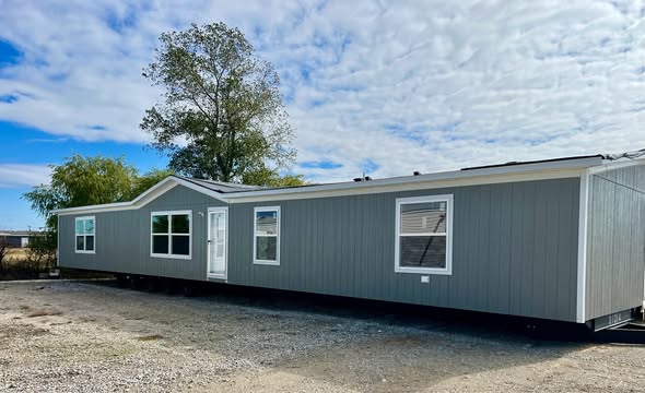 A modern, gray mobile home with white trim sits on gravel. Large windows and trees in the background reflect a clear sky, conveying a serene setting.