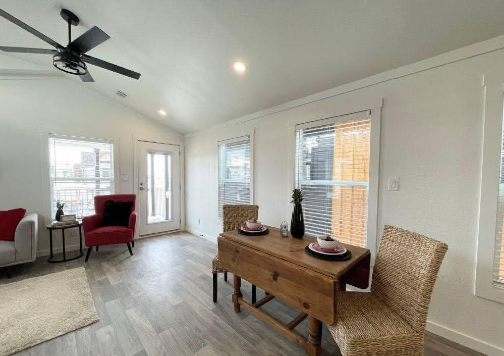 A bright living space with light wood floors and white walls. A ceiling fan, red chair, and a wooden table set for two add a cozy, modern feel.