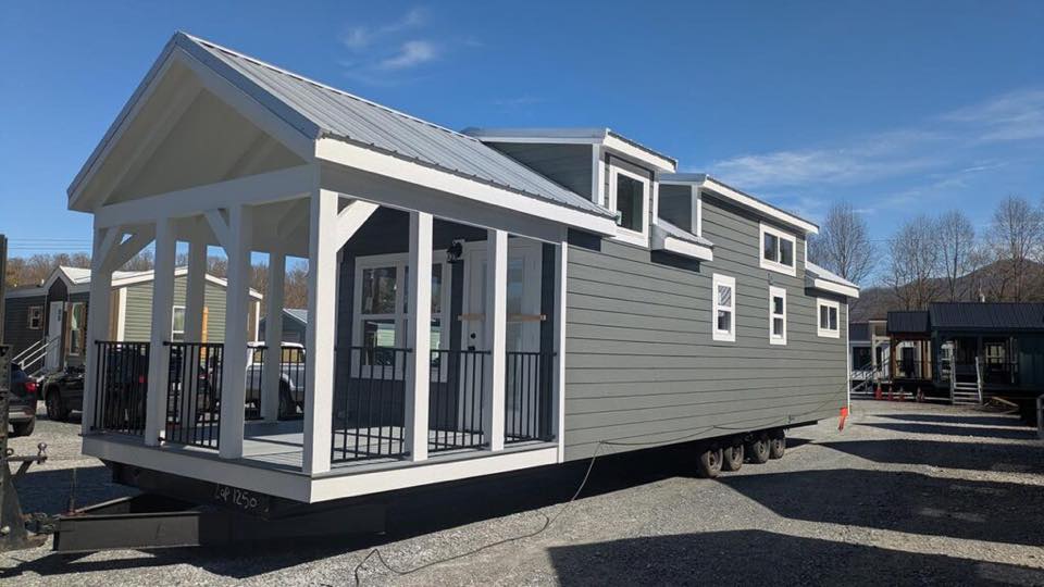 A modern tiny house on wheels with gray siding, white trim, and a spacious porch. The clear sky and other tiny homes in the background suggest a calm day.