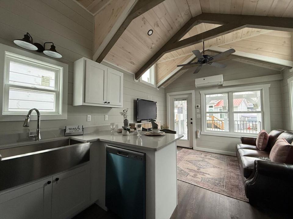 Cozy living area with high vaulted ceiling and exposed beams. Features a kitchenette with white cabinets, a leather couch, and large windows offering ample natural light.
