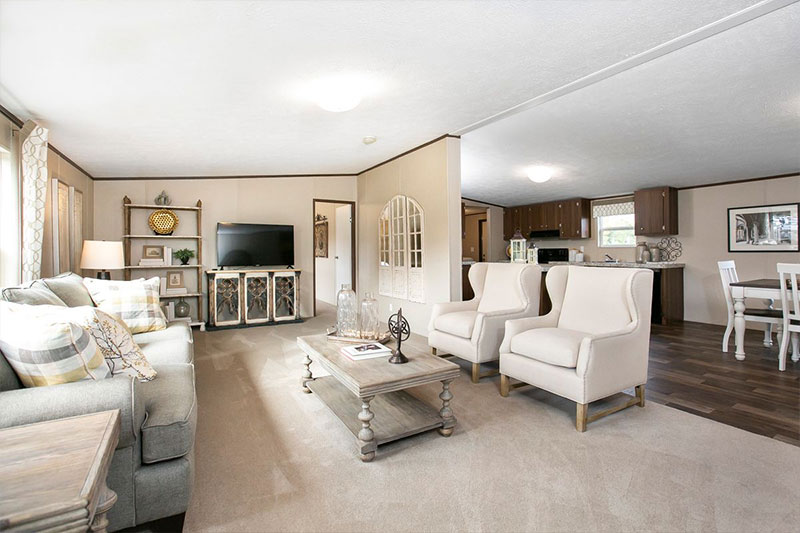 Bright living room with beige walls and carpet, featuring a gray sofa, two white armchairs, and wooden coffee table. TV and a dining area visible. Cozy and inviting.