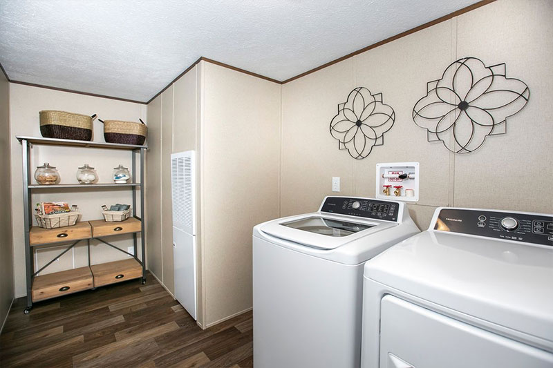 Laundry room with beige walls, featuring a washer and dryer, decorative metal wall art, and a shelving unit with wicker baskets and storage jars. Warm and organized.