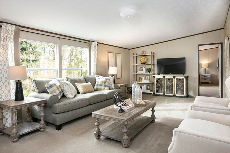 Bright living room with a large gray sofa, plaid pillows, two beige armchairs, wooden coffee table, and a TV on a decorative cabinet. Large windows on the left.