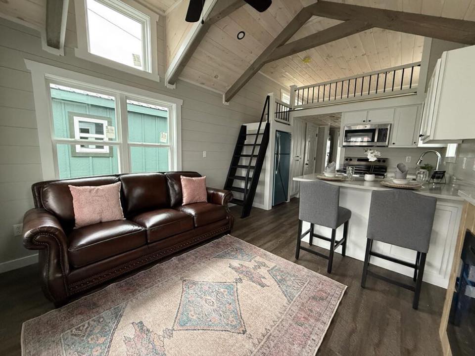 Cozy tiny home interior with a vaulted ceiling. Features a brown leather sofa, pastel rug, modern kitchen, and loft with a ladder. Warm and inviting.
