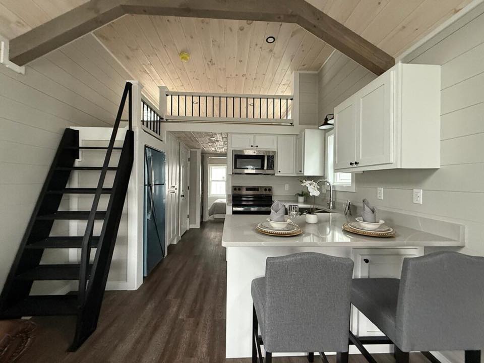 Cozy modern kitchen with high vaulted ceiling, wooden beams, and a loft. Features dark wood floors, white cabinets, and two bar stools at a counter.